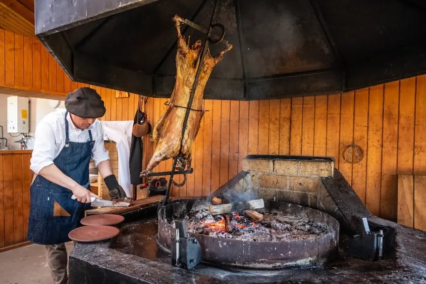 A man in a chef’s apron slices lamb meat next to a fire where a whole lamb is being roasted.